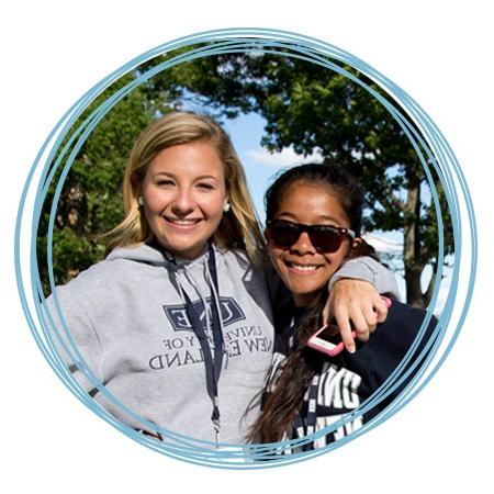 Two smiling students in U N E sweatshirts stand outdoors together on the Biddeford Campus